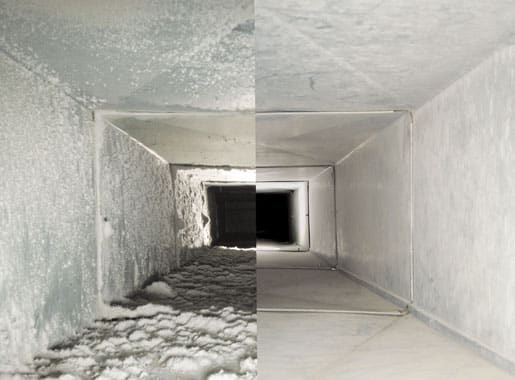 Interior view of a commercial air duct or HVAC ventilation system showing significant dust and debris accumulation on all surfaces, including walls, ceiling, and floor. The rectangular tunnel extends toward a window opening at the far end, with stark lighting revealing the poor condition and lack of maintenance. This image illustrates the importance of regular duct cleaning for air quality and system efficiency.