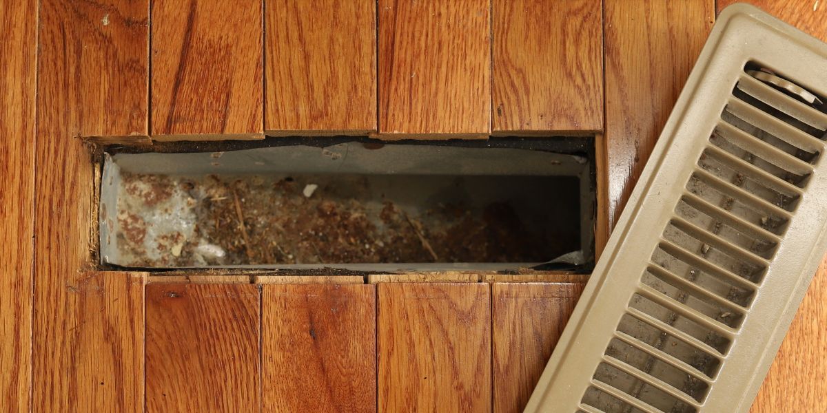 A wooden floor with visible rust and debris accumulation inside a dark rectangular floor vent or duct opening on the left, next to a cream-colored air return vent on the right. The wood flooring shows natural grain patterns and aging, while the metal vent interior displays significant rust staining and dust buildup, indicating neglect and poor maintenance of the HVAC system.