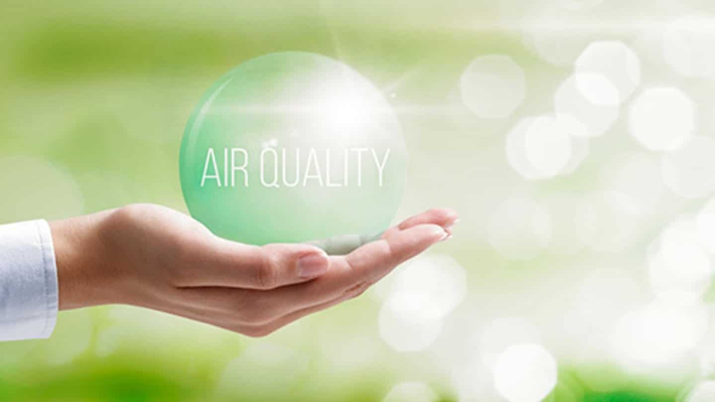 A hand emerging from a white sleeve holds a translucent green sphere with "AIR QUALITY" text displayed on it against a soft, blurred background of green foliage and white bokeh lights. The image symbolizes environmental awareness and the importance of clean air, using the globe to represent Earth and sustainability. The gentle, nature-inspired composition conveys a message of care and responsibility toward air quality and environmental protection.
