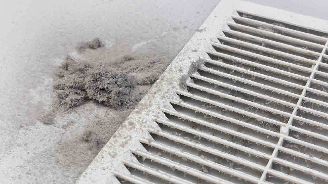 A close-up view of a white or light-colored air vent or register on a ceiling or wall, with a visible accumulation of dust and debris on the surrounding surface. The dust buildup appears as a gray, fuzzy cluster next to the vent's horizontal slats, indicating poor air filter maintenance or prolonged lack of cleaning in the HVAC system.