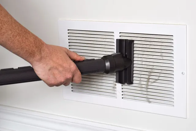A person's hand holding a black cleaning brush tool against a white wall-mounted air vent with horizontal slats. The brush appears to be designed specifically for cleaning the narrow spaces between vent louvers, demonstrating maintenance of home HVAC systems.