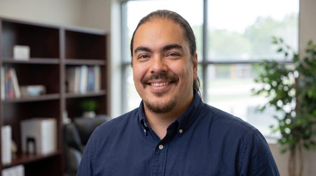 A smiling man with long dark hair and a mustache wearing a navy blue button-up shirt stands in a bright, modern office space. Behind him are wooden shelving units filled with books and decorative items, along with potted plants near large windows that overlook a green outdoor landscape. The setting suggests a professional workspace with a welcoming, friendly atmosphere.