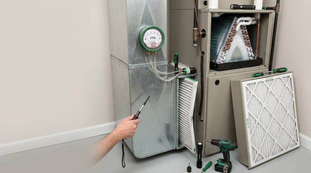 A technician holds a digital thermometer probe while performing HVAC maintenance on residential heating and cooling equipment. The setup includes a furnace unit with a pressure gauge reading 0.90, exposed ductwork, air filters, and various tools including a cordless drill positioned on the floor. The work appears to be part of a routine inspection or service call in a home's mechanical room.