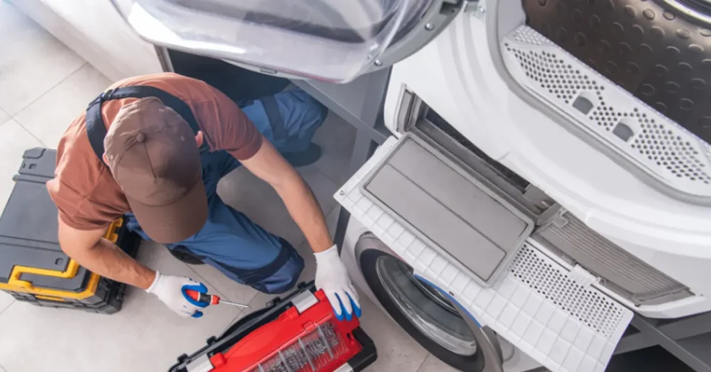 A technician in an orange shirt and blue pants performs maintenance or repair work on laundry appliances, kneeling on a white floor surrounded by a washing machine, dryer, and various tools including a red toolbox and yellow level. The perspective is from above, showing the technician focused on their work as they service the stacked white laundry equipment in what appears to be a residential laundry room.
