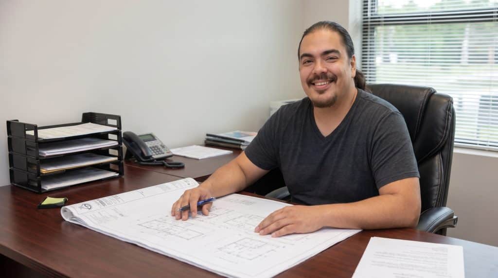 A professional man with long dark hair and a mustache sits at a wooden desk in a modern office, smiling at the camera while reviewing architectural blueprints. He wears a dark gray v-neck t-shirt and is positioned in front of a black leather office chair with windows visible behind him. The desk is organized with a document tray, phone, papers, and sticky notes, suggesting an active work environment in architecture or engineering.