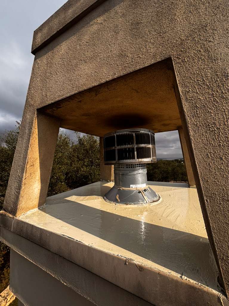 A concrete brutalist structure with a large open window frame reveals a cylindrical metal chimney or ventilation pipe inside. The structure appears to be an industrial or architectural monument set in a rural landscape with trees visible in the background under an overcast sky. Sunlight casts shadows across the interior concrete floor, highlighting the stark, geometric design of the building.
