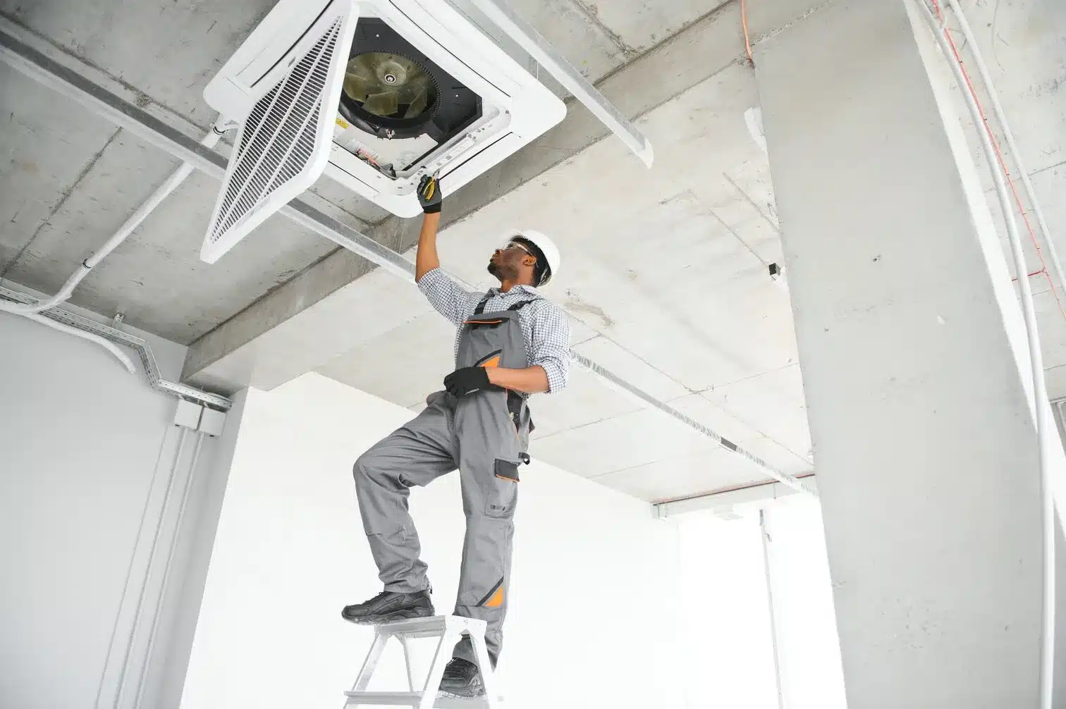 A technician in gray work overalls and safety glasses stands on a white ladder, reaching up to service a ceiling-mounted HVAC unit in an unfinished interior space. He is wearing work gloves and appears to be performing maintenance or installation work on the air conditioning system. The room has exposed concrete ceiling, white walls, and electrical conduits visible overhead.