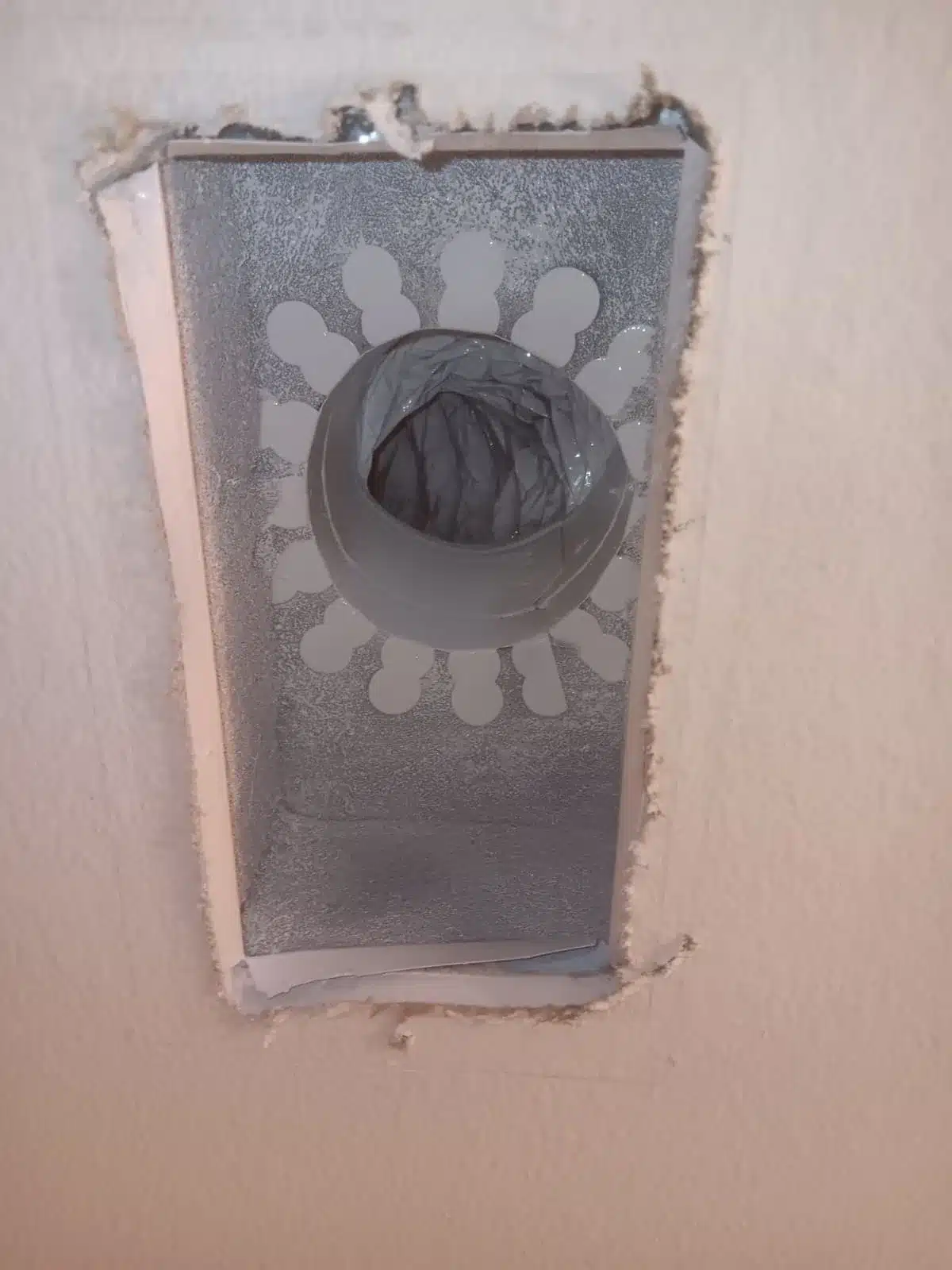 Rectangular metal duct component exposed in a drywall opening, with a round central connection and dark interior.